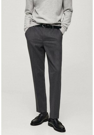 Homme portant un pantalon gris foncé sur mesure, une ceinture noire, des mocassins en cuir noir et un pull gris clair, les mains dans les poches.