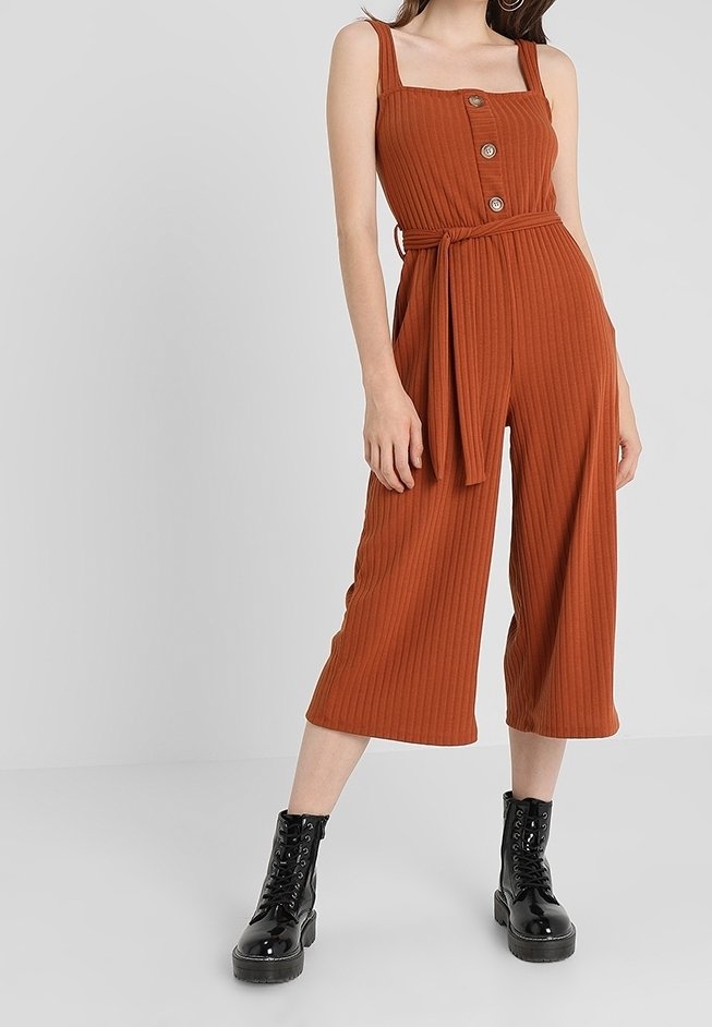 Femme portant une combinaison sans manches côtelée couleur rouille avec des boutons sur le devant et une ceinture, associée à des bottines noires lacées, debout devant un fond gris.