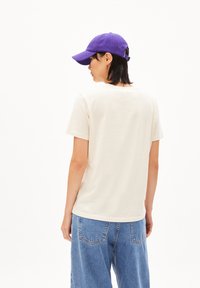 Beige Baumwoll-T-Shirt mit Rundhalsausschnitt; kombiniert mit hellblauen Jeans. Eine lila Mütze wird getragen, die ein einfaches Design zeigt.