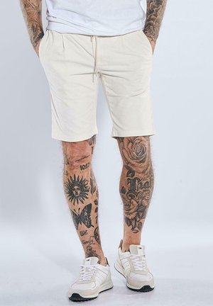 Man wearing cream shorts and white sneakers, with tattooed legs featuring sun, butterfly, roses, and various text designs. Hands in pockets.