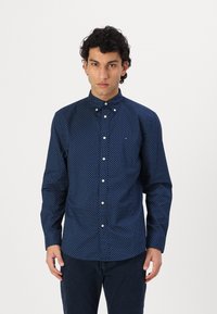 Navy blue button-up shirt with white polka dots, long sleeves, and a button-down collar. Notable logo on the chest, smooth fabric texture.