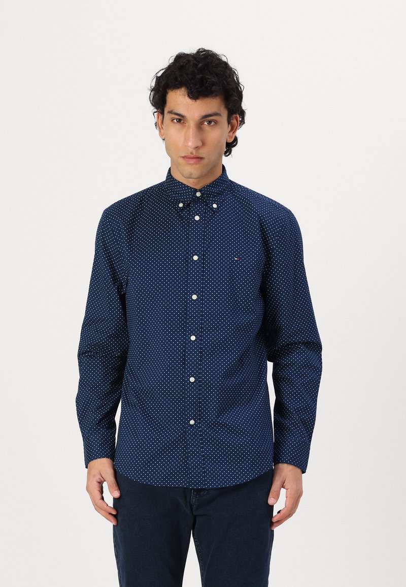 Navy blue button-up shirt with white polka dots, long sleeves, and a button-down collar. Notable logo on the chest, smooth fabric texture.