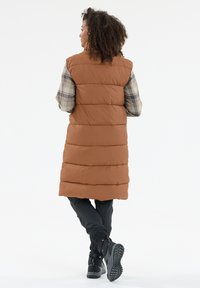 Bruine puffer vest met verticale naden, knielange bedekking en een gewatteerde textuur. Gedragen over een geruit shirt, gecombineerd met zwarte broek en laarzen.