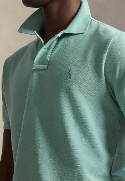 Άνδρας με ανοιχτοπράσινο πόλο πουκάμισο με ανασηκωμένο γιακά και μικρό κεντητό μπλε λογότυπο στο στήθος.