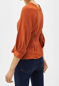 Kvinde iført en struktureret burnt orange bluse med trekvart lange pufærmer, parret med mørkeblå tætsiddende jeans, stående mod en ensfarvet baggrund.