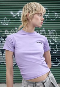 Vijolična oprijeta kratka majica s kratkimi rokavi, z črno nalepko "DICKIES" na prsih. Material je videti gladek, dopolnjen s sivimi hlačami.