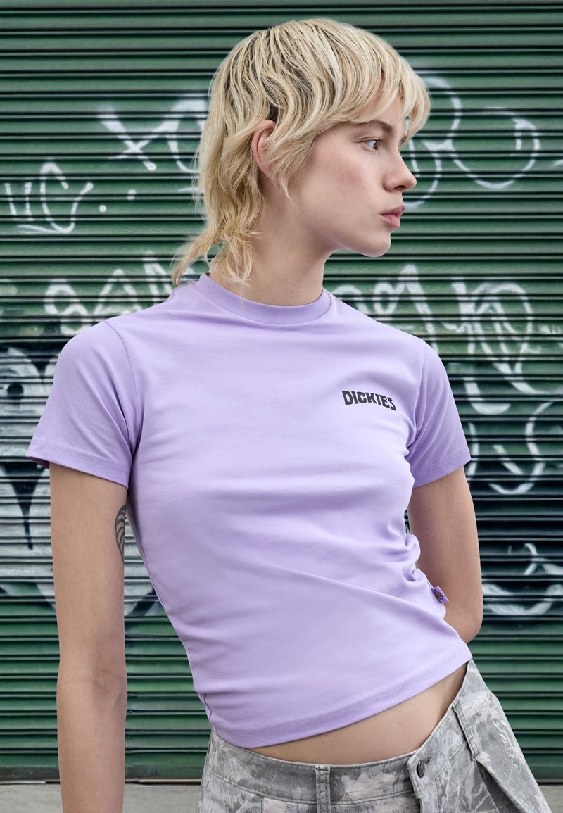 Vijolična oprijeta kratka majica s kratkimi rokavi, z črno nalepko "DICKIES" na prsih. Material je videti gladek, dopolnjen s sivimi hlačami.