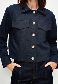 Chaqueta recortada de mezclilla azul oscura con dos bolsillos en el pecho, botones dorados y un cuello estructurado. Textura suave con detalles de costura visibles.