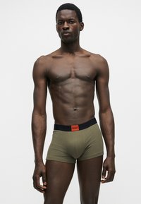 Modèle masculin à la peau foncée portant un boxer vert olive avec une ceinture noire et un logo Hugo rouge, debout sur un fond clair.
