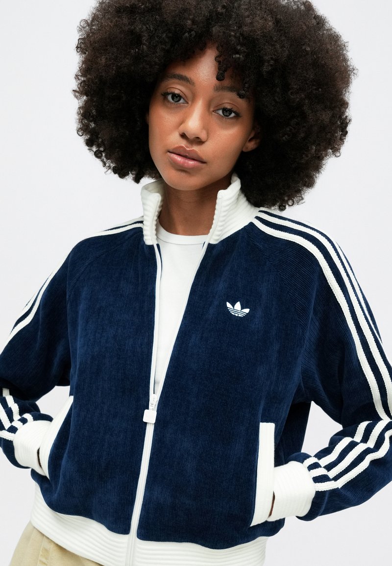 Veste zippée en velours côtelé bleu marine avec col et poignets côtelés blancs, comportant trois bandes blanches sur chaque manche et un petit logo brodé.