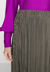 Kvinna som bär en långärmad lila blus och en höga midjan veckad olivgrön kjol, med handen avslappnad vid sin sida.