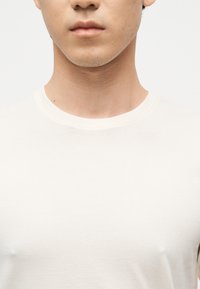 Νέος άνδρας που φοράει ένα απλό λευκό μπλουζάκι με στρογγυλή λαιμόκοψη, εμφανίζοντας το λαιμό και το πάνω μέρος του στήθους του, μπροστά σε ουδέτερο φόντο.