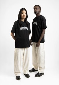 Black graphic t-shirts with white text, oversized fit, paired with wide creamy corduroy pants and black footwear, against a plain background.