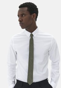 Chemise blanche à boutons, cravate verte texturée, pantalon noir, portée par une personne avec des cheveux courts et bouclés. Lignes simples et épurées, avec un col visible.