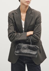 Zwarte handgeweven handtas met een gestructureerde afwerking, ritsluiting en twee korte handvatten, vastgehouden door een persoon in een grijze blazer en een witte top.