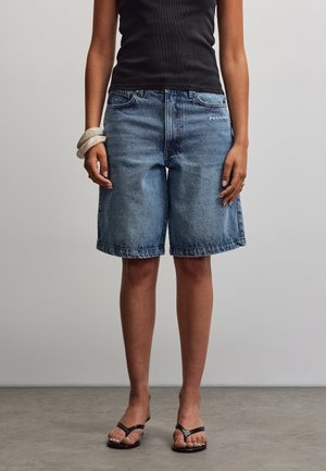 Persoană purtând pantaloni scurți din denim albastru până la genunchi, top negru fără mâneci, sandale negre și brățări albe la încheietura mâinii stângi, stând pe podea gri.