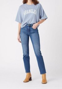 Camiseta oversized de color azul claro con estampado "WRANGLER", combinada con jeans rectos de color azul oscuro y botas de tobillo beige con tacón cuadrado.