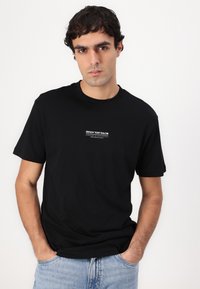 Sort bomulds T-shirt med rund hals og korte ærmer, prydet med et hvidt grafisk design på forsiden. Båret med lyse denim jeans.