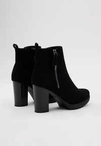 Botas de tobillo de ante negro con un tacón grueso, cierre lateral y suela texturizada. El diseño cuenta con un acabado suave y punta redondeada.