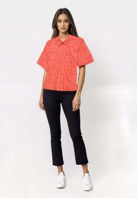 Korallfarget kortermet skjorte med utskårent mønster og knapper foran, kombinert med svarte capribukser og hvite joggesko.