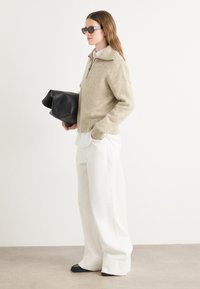 Suéter bege de malha com colarinho de meia zíper, calças brancas de corte largo e bolsa preta de couro. Tecido texturizado volumoso. Óculos de sol quadrados pretos.