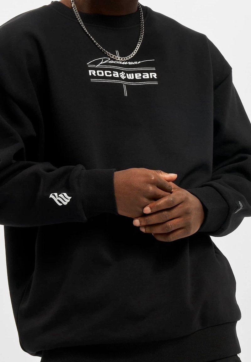 Sweatshirt noir avec un col rond et des poignets côtelés, arborant un logo "ROCA WEAR" blanc sur le devant et un accent à motifs sur la manche.
