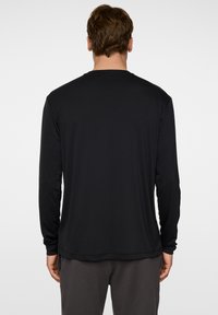 Musta pitkähihainen paita, joka on valmistettu kevyestä ja sileästä kankaasta. Ominaisuuksiin kuuluu pyöreä kaula-aukko ja ommeltu helma. Minimalistinen muotoilu ilman näkyviä logoja.