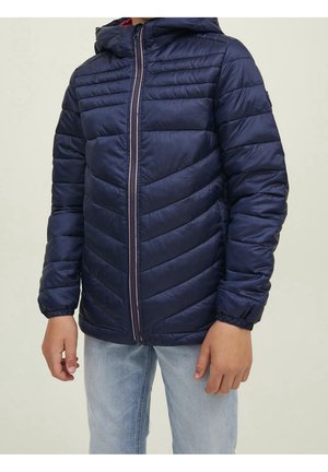 Bambino con giacca imbottita blu navy con cappuccio e cerniera frontale, e jeans azzurri, in piedi con le braccia rilassate.