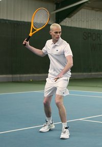 Tennisspieler in einem weißen Polo-Shirt und Shorts, der einen orangenen Schläger hält. Der Platz hat eine blaue Oberfläche und einen grünen Hintergrund, mit einem Netz im Hintergrund.