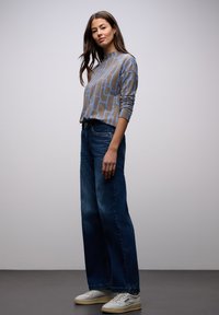 Suéter de color marrón claro con patrón geométrico azul, combinado con jeans de pierna ancha azul oscuro y zapatillas blancas. Textura suave, ajuste relajado.