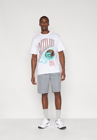 Λευκό γραφικό μπλουζάκι με το κείμενο "Memphis Grizzlies" και σχέδιο αρκούδας, συνδυασμένο με γκρι σορτς και λευκά αθλητικά παπούτσια.