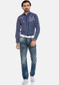 Marineblauwe geblokte overhemd met rode en witte borduursels, gecombineerd met vervaagde blauwe jeans en een witte riem, gedragen met witte sneakers.