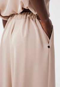 Pantalons beige, en tissu soyeux et texturé, avec une coupe ample, dotés d'une taille froncée et de poches latérales fermées par de petits boutons noirs.