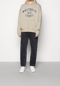 Beige hoodie met "WRSTBHVR STUDIO"-opdruk, gecombineerd met donkergrijze losse jeans en witte sneakers.