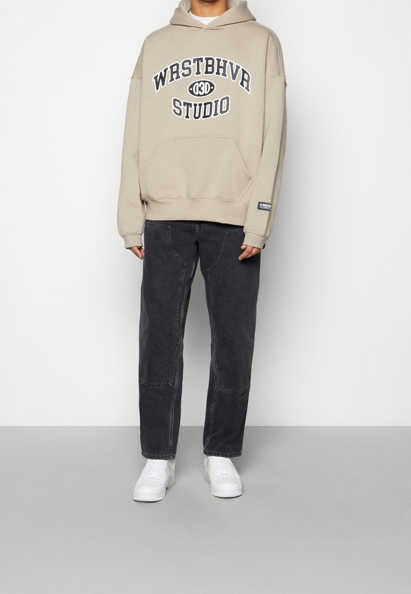 Beige hoodie met "WRSTBHVR STUDIO"-opdruk, gecombineerd met donkergrijze losse jeans en witte sneakers.