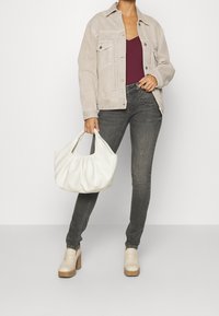 Frau in beiger Jacke, burgunderrotem Oberteil, grauen Skinny-Jeans und beigen Plateauschuhen, die eine weiße, weiche Leder-Handtasche vor einem schlichten Hintergrund hält.