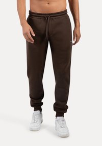 Brune bomuldsjoggingbukser med elastisk talje og snor, der har sidelommer og en tætsiddende benpasform. Sammen med hvide sneakers.