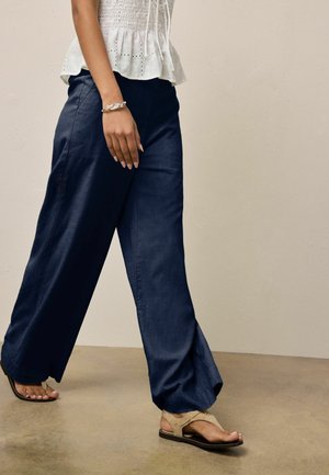 Woman wearing navy wide-leg trousers, white eyelet top, beige sandals, and silver twisted bracelet walking on tan floor against beige wall.