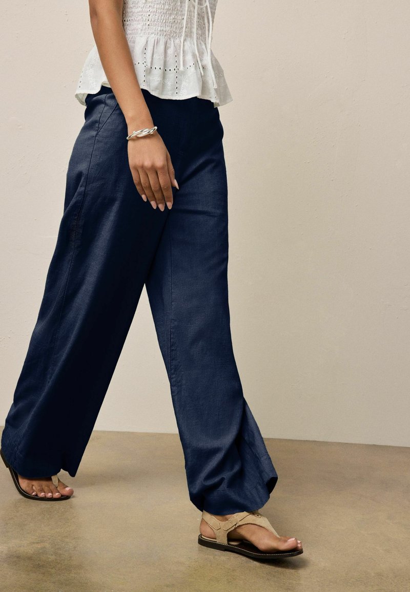Femme portant un pantalon large bleu marine, un haut blanc en broderie anglaise, des sandales beiges et un bracelet torsadé argenté, marchant sur un sol beige devant un mur beige.