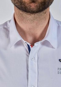 Homme barbu portant une chemise blanche à col avec des bordures rouge, blanche et bleue à l'intérieur du col, un texte brodé partiellement visible.