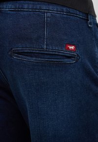 Dunkelblaue Jeans aus Denim mit einer hinteren Tasche, die mit einem kleinen roten Etikett mit weißen Pferde-Design versehen ist. Der Stoff hat eine glatte Textur.