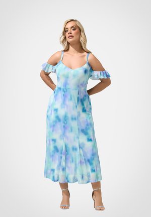 Femme debout, mains dans le dos, portant une robe midi aquarelle bleue et violette à épaules dénudées et des sandales argentées à talons hauts.