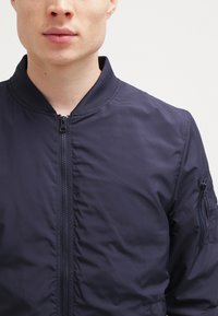 Jachetă de bomber marină, cu fermoar frontal, guler și manșete ribbate. Include buzunare laterale și un mic buzunar cu fermoar pe mânecă. Textură netedă.