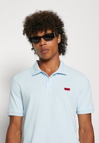 Ljusblå pikétröja med ett rött logotypmärke på vänster bröst. Modellen bär svarta solglasögon. Tyget verkar slätt med korta ärmar.