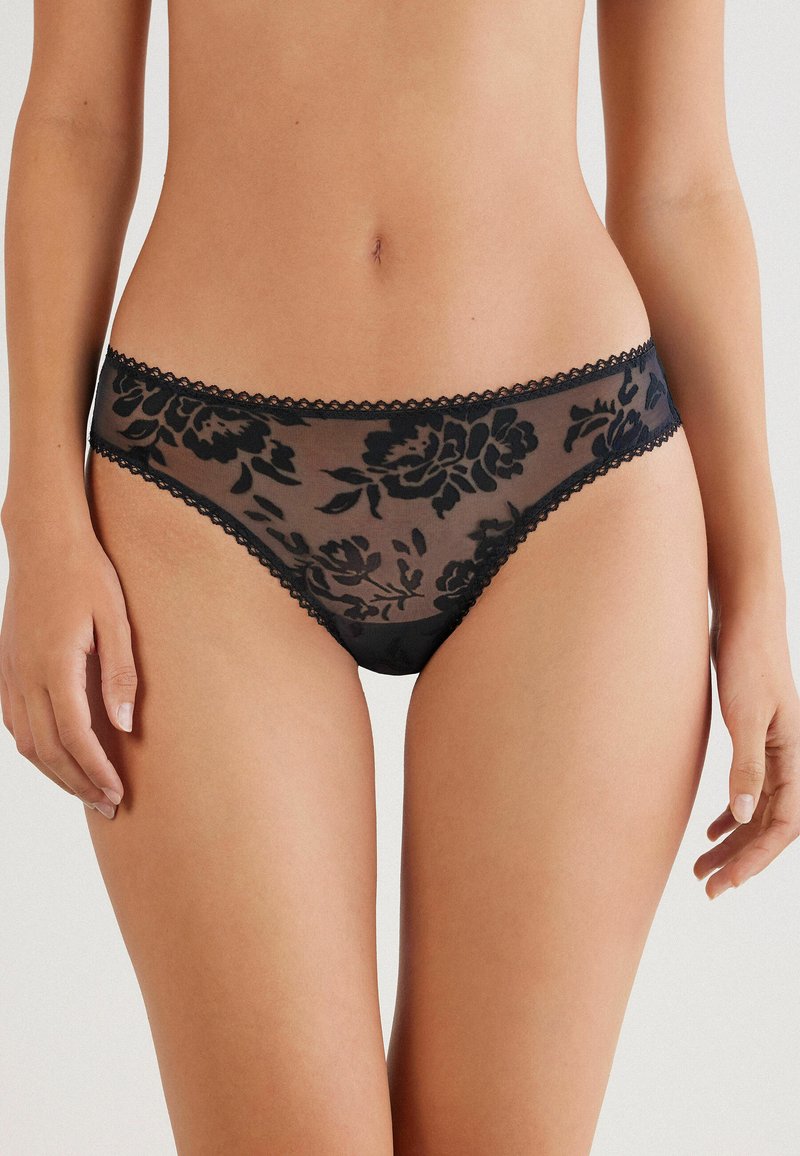 Svart genomskinlig spetsstring med blommönster, vågig kant längs midjebandet och en mjuk textur för en delikat design.