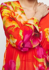 Bloemenjurk met levendig rode stof en roze en gele bloemendessins, lange mouwen met gathered cuffs en een V-halsontwerp.