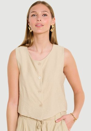 Junge Frau trägt ein ärmelloses beigefarbenes, mit Knöpfen versehenes Oberteil und passende Hose mit goldenen Ohrringen und Armband, Hand in der Tasche ruhend.