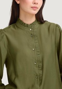 Blusa verde oliva con colletto alzato, rifinitura a rouches verticale e chiusura a bottoni. Tessuto morbido con una superficie liscia e leggermente lucida.