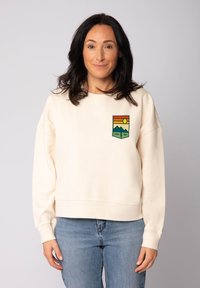 Vrouw met donker haar draagt een crèmekleurige sweatshirt met een kleurrijke berg- en zonpatch met het label "Good Vibe" en lichte blauwe jeans, staand tegen een effen achtergrond.