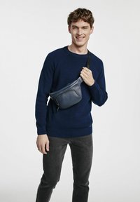 Niebieska skórzana torebka typu crossbody z kieszonką na zamek i regulowanym paskiem, noszona na ciemnym swetrze i szarych dżinsach, na jednolitym tle.